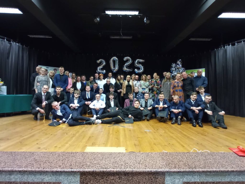 Duża grupa ludzi, w tym studenci w garniturach i dorośli, pozują na scenie z balonami 2025 w tle, sugerując wydarzenie z okazji ukończenia szkoły lub uroczystości. Niektórzy ludzie siedzą, inni stoją, wszyscy się uśmiechają.
