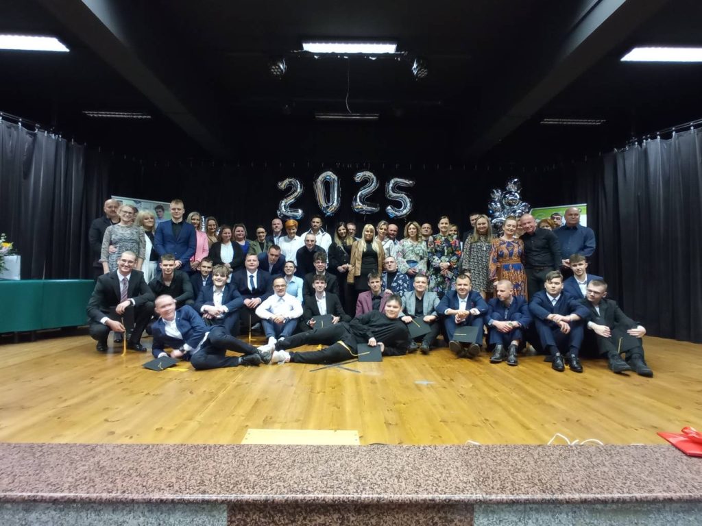 Duża grupa osób, w tym uczniowie w garniturach i dorośli, pozuje razem na scenie, a w tle widać dekoracje z balonów z napisem „2025”, co sugeruje uroczystość ukończenia szkoły lub klasową uroczystość.