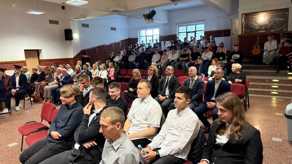 Grupa ludzi, w większości ubranych formalnie, siedzi w rzędach krzeseł w pomieszczeniu z drewnianymi panelami na ścianach, uczestnicząc w wydarzeniu lub ceremonii w pomieszczeniu. Publiczność obejmuje zarówno młodych dorosłych, jak i osoby starsze.