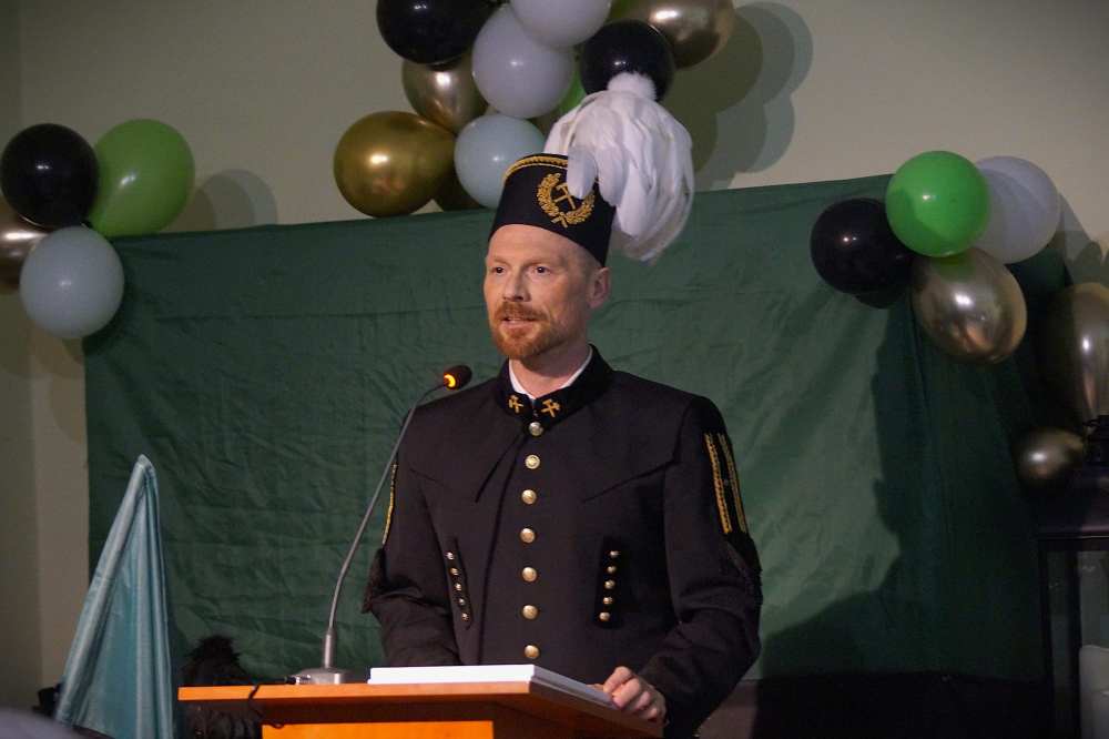 Mężczyzna w tradycyjnym czarnym mundurze ze złotymi guzikami i kapeluszu z piórami przemawia z podium. Za nim znajdują się białe, czarne i złote balony oraz zielone tło z tkaniny.