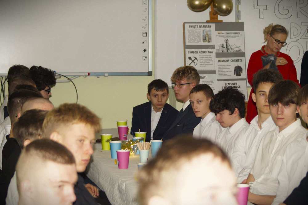 Grupa formalnie ubranych młodych ludzi siedzi wokół długiego stołu z kolorowymi kubkami i przekąskami. W tle kobieta stoi w pobliżu tablicy i plakatów. Atmosfera wydaje się poważna i uważna.