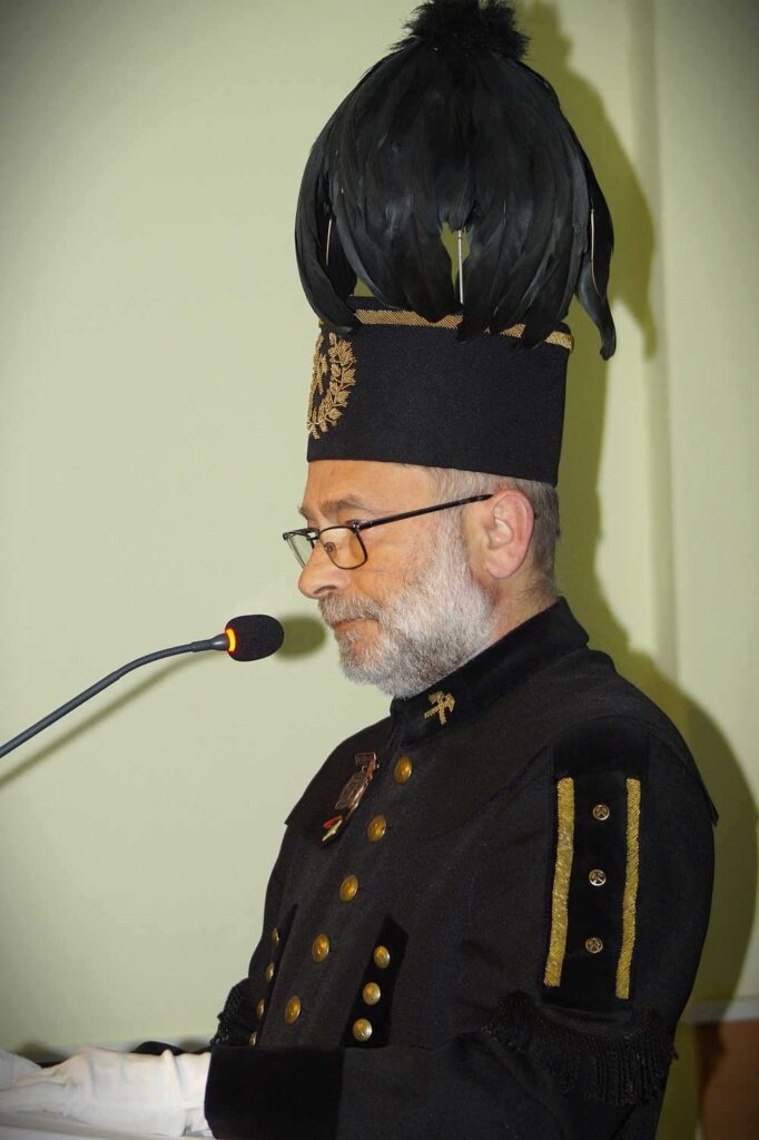 Mężczyzna w okularach i z siwą brodą, ubrany w czarny ceremonialny mundur ze złotymi akcentami i wysoki kapelusz z piórami, przemawia do mikrofonu na podium.