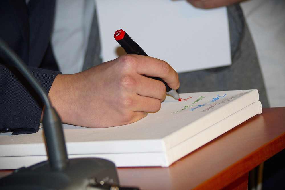 Osoba pisze kolorowy tekst na białym papierze czarnym i czerwonym markerem, siedząc przy biurku. W tle widoczny jest mikrofon i inna osoba trzymająca dokumenty.