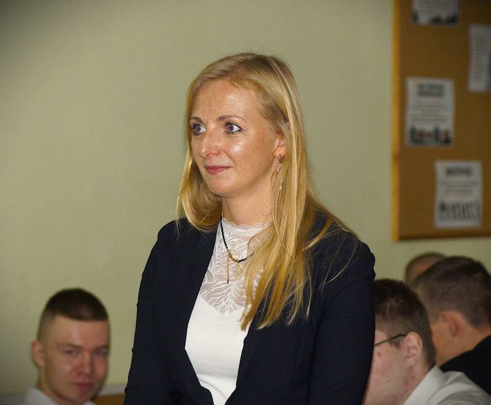 Kobieta z długimi blond włosami, ubrana w ciemną marynarkę na białym topie, stoi w pomieszczeniu. W tle siedzi kilka osób, a za nimi na ścianie widoczna jest tablica ogłoszeń.