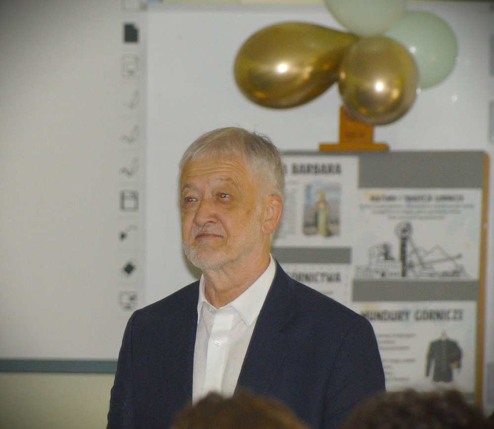 Starszy mężczyzna z siwymi włosami i brodą, ubrany w białą koszulę i ciemną marynarkę, stoi w pomieszczeniu przed tablicą i plakatami. W tle widoczne są złote i białe balony.