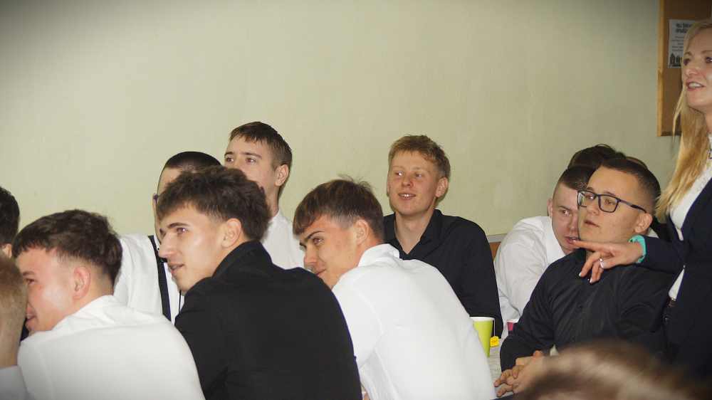 Grupa młodych ludzi, głównie mężczyzn, ubranych w białe i czarne koszule, siedzi blisko siebie przy stołach, wyglądając na zaangażowanych i uśmiechniętych. Kobieta po prawej stronie stoi i wydaje się mówić lub wskazywać.