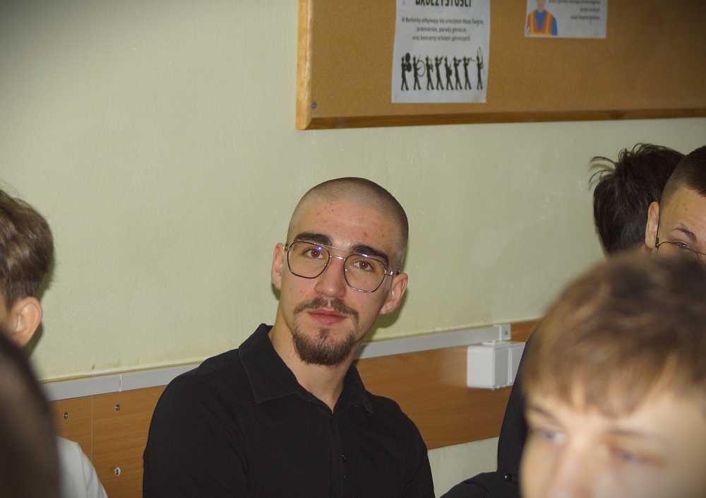 Młody mężczyzna z ogoloną głową, okularami i kozią bródką siedzi w pomieszczeniu i patrzy w kamerę. Znajduje się w klasie z innymi ludźmi wokół i tablicą ogłoszeń na ścianie za nim.