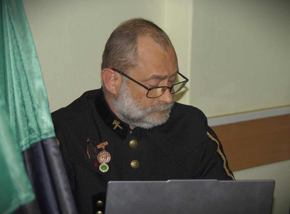 Mężczyzna w okularach z siwą brodą, ubrany w ciemną marynarkę ze szpilkami i guzikami, siedzi przy biurku i patrzy w dół na laptopa. Zielono-czarna tkanina jest częściowo widoczna po lewej stronie.