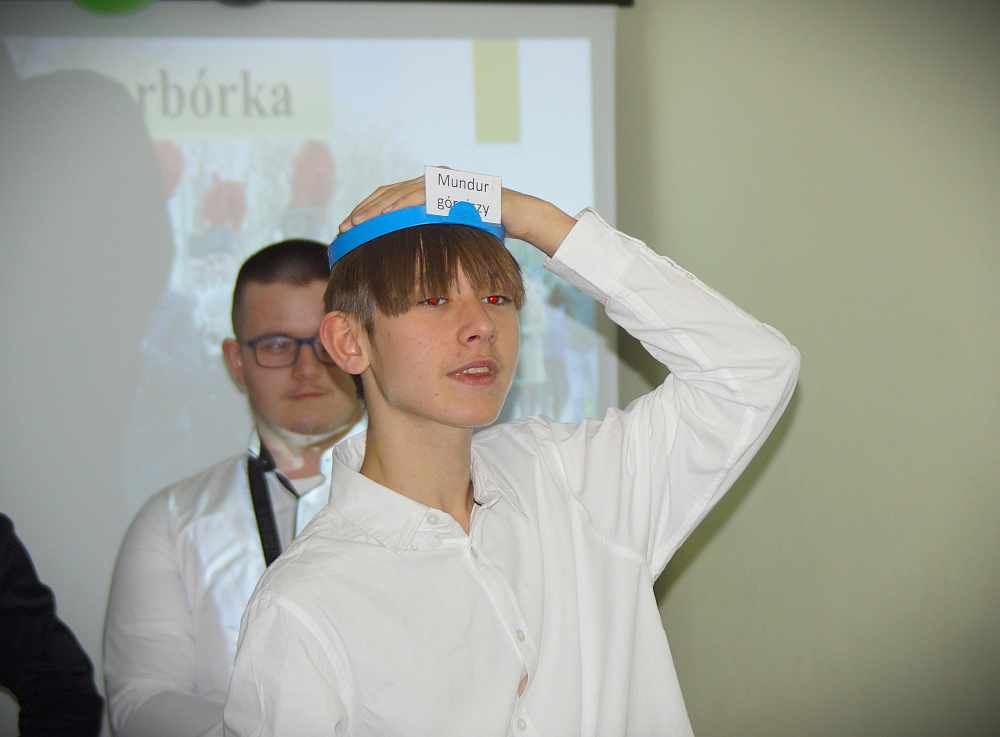 Chłopiec w białej koszuli trzyma niebieską opaskę z kartą na czole, grając w zgadywankę. W tle stoi inna osoba w okularach i białej koszuli, a za nią wyświetlany jest ekran.