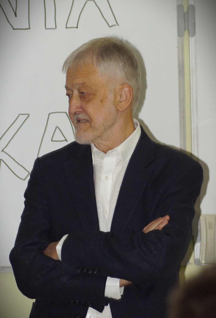 Starszy mężczyzna z krótkimi siwymi włosami i brodą, ubrany w ciemną marynarkę i białą koszulę, stoi ze skrzyżowanymi rękami, patrząc w bok przed tablicą z dużymi, zarysowanymi literami.