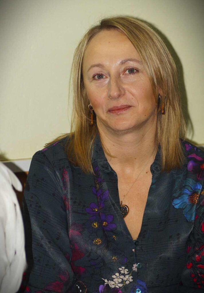 Kobieta o prostych blond włosach ubrana w ciemną bluzkę w kwiaty patrzy w kamerę z lekkim uśmiechem. Siedzi w pomieszczeniu przy jasnej ścianie.