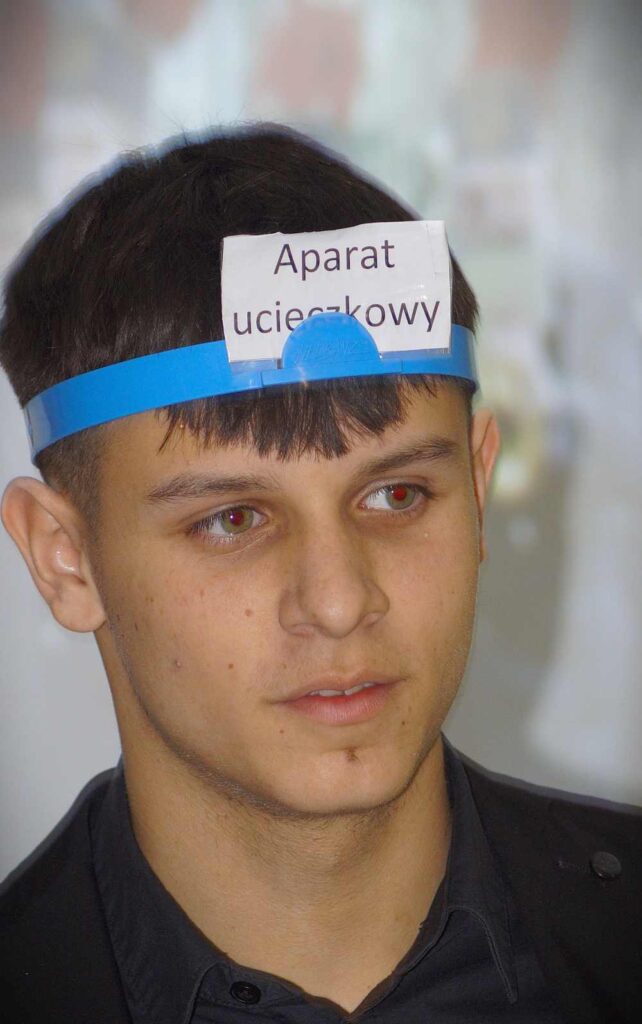 Młody mężczyzna o krótkich ciemnych włosach nosi na czole niebieską opaskę z papierową etykietą Aparat ucieczkowy. Patrzy lekko w bok z neutralnym wyrazem twarzy.