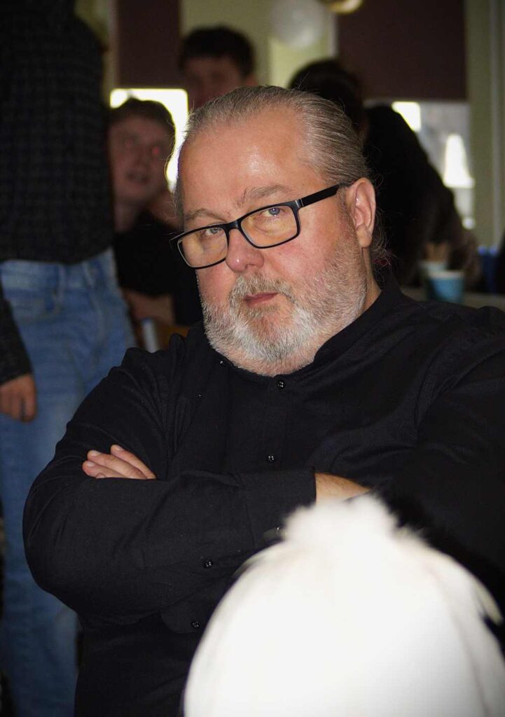 Mężczyzna z siwymi włosami, brodą i w okularach, ubrany w czarną koszulę, siedzi ze skrzyżowanymi ramionami. Patrzy bezpośrednio w kamerę z poważnym wyrazem twarzy. Za nim znajdują się ludzie i rozmyte tło.