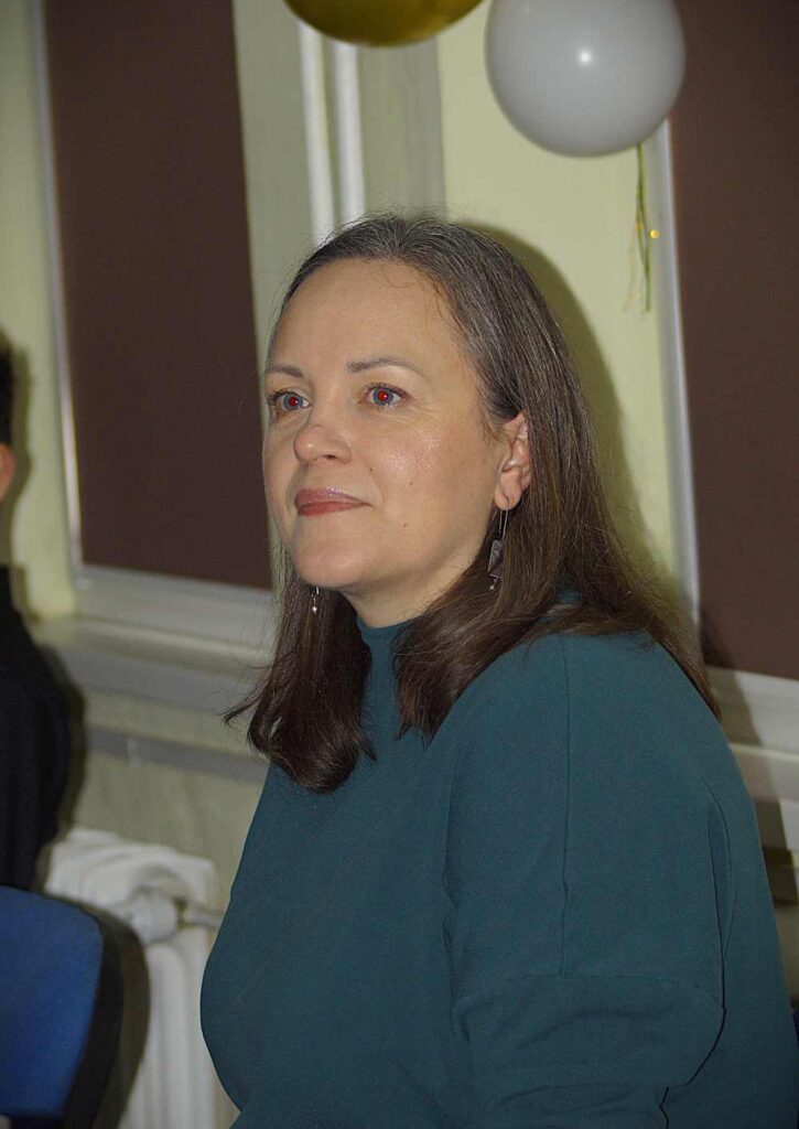 Kobieta z prostymi brązowymi włosami ubrana w zielony top siedzi w pomieszczeniu w pobliżu balonów, patrząc lekko w bok. Tło stanowią brązowe panele ścienne i biały grzejnik.