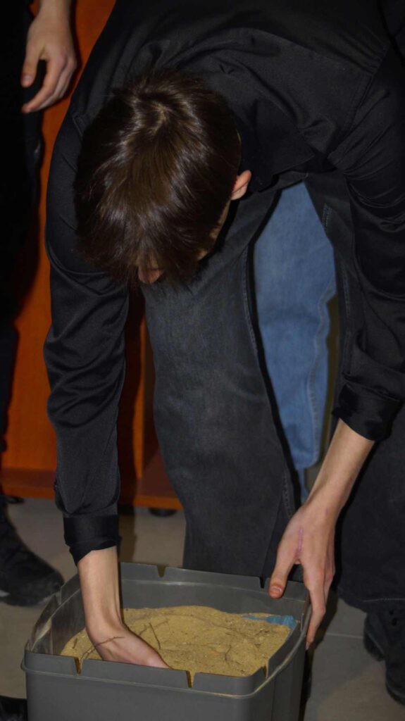 Osoba w ciemnym ubraniu pochyla się i sięga do szarego pojemnika wypełnionego piaskiem lub podobnym materiałem. Inna osoba stoi w pobliżu, częściowo widoczna w tle.