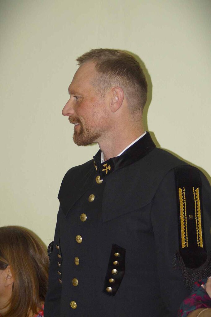 Mężczyzna z krótkimi włosami i brodą ubrany jest w ciemny, zapinany na guziki mundur ze złotymi akcentami i haftami, stojąc profilem na jasnym tle. Inna osoba jest częściowo widoczna, siedzi obok niego.