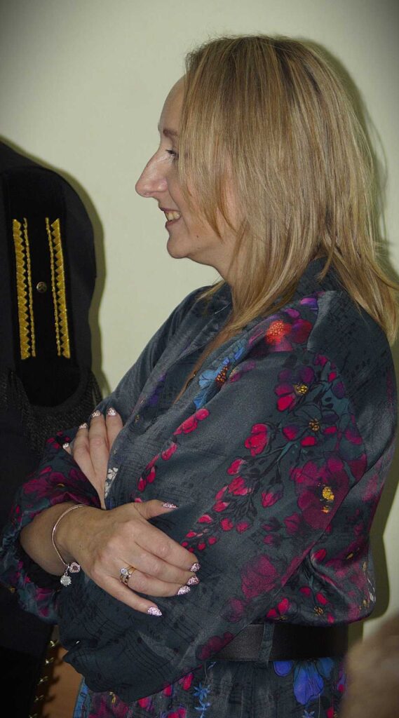 Kobieta o blond włosach stoi z profilu ze skrzyżowanymi ramionami, ubrana w ciemną sukienkę w kwiatowe wzory. Obok niej częściowo widoczna jest inna osoba w ciemnym stroju.