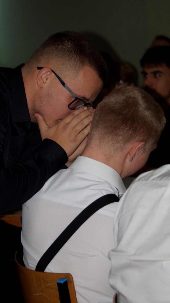 Młody mężczyzna w okularach pochyla się, by szepnąć do ucha innemu młodemu mężczyźnie. Obaj siedzą, a scena wydaje się być w pomieszczeniu z innymi ludźmi w tle.