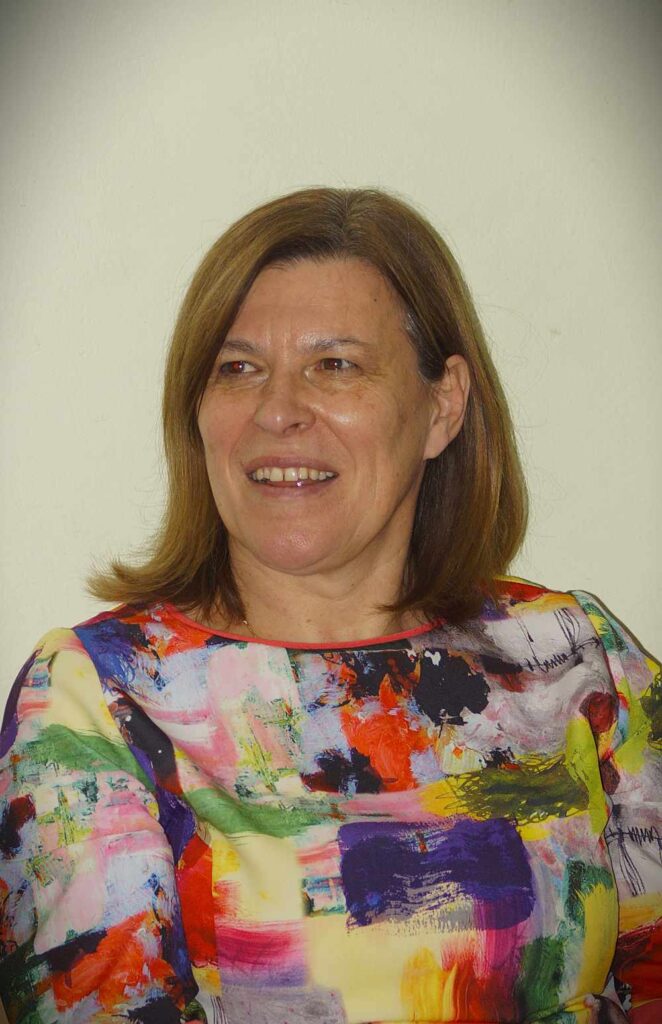 Kobieta z prostymi brązowymi włosami uśmiecha się lekko, ubrana w kolorowy top z abstrakcyjnym wzorem, siedząca przed prostym, jasnym tłem.