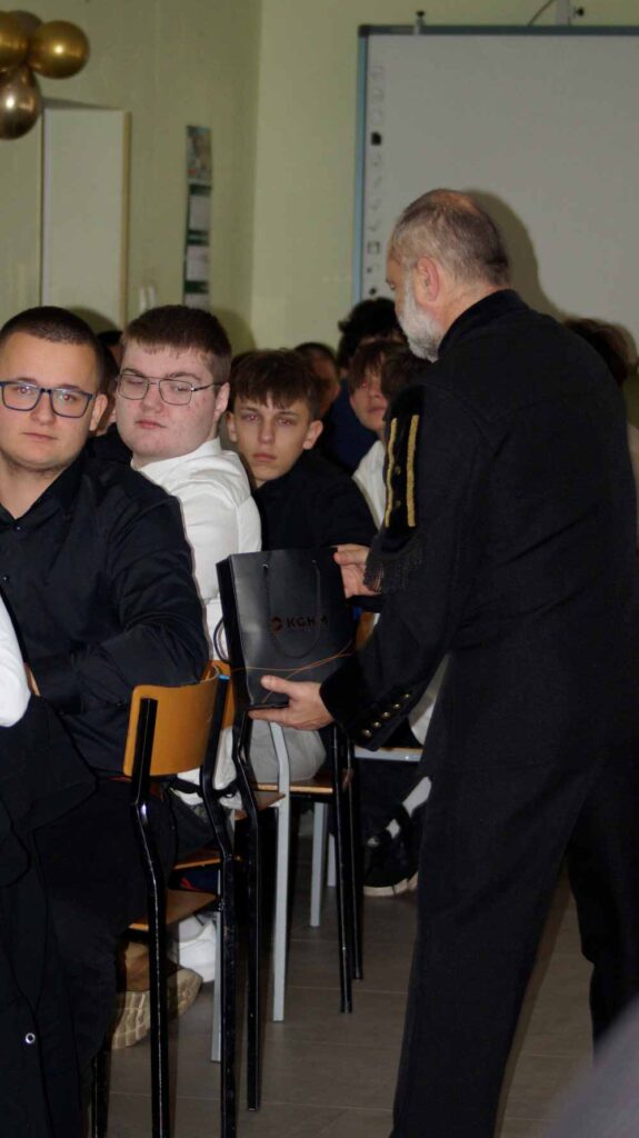 Mężczyzna z siwymi włosami i brodą, ubrany w ciemne ubranie, wręcza czarną teczkę siedzącemu młodemu mężczyźnie w klasie wypełnionej uczniami. Inni uczniowie siedzą przy ławkach, obserwując interakcję.