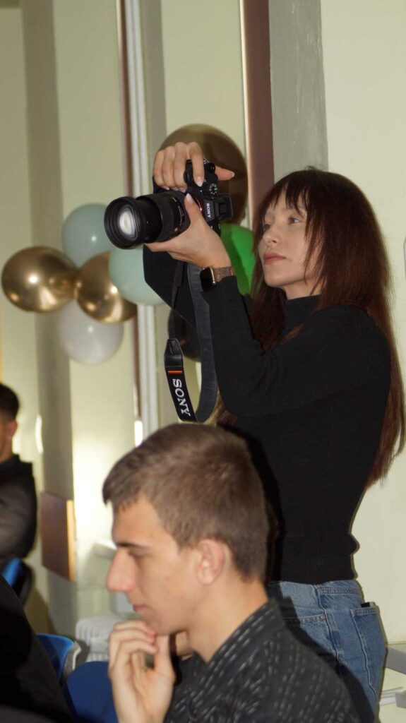 Kobieta stoi trzymając aparat Sony, robiąc zdjęcie na imprezie w pomieszczeniu ze złotymi, białymi i zielonymi balonami w tle. Siedząca osoba jest zamazana na pierwszym planie.