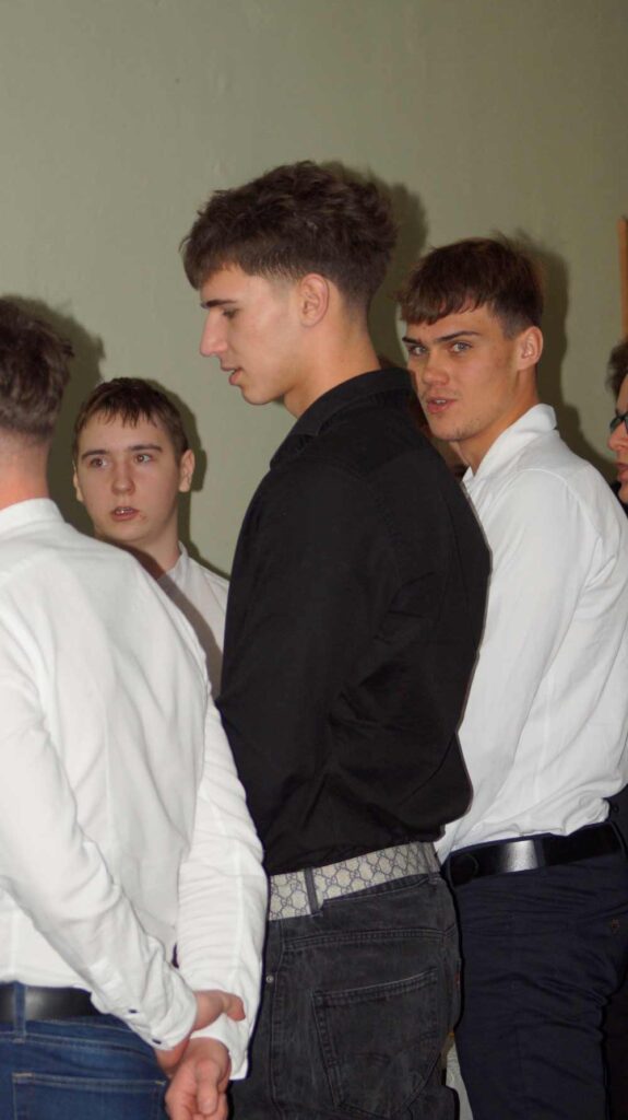 Czterech młodych mężczyzn stoi w pomieszczeniu, zwróceni do siebie bokiem i rozmawiają. Trzech z nich ma na sobie białe koszule, a jeden czarną. Tło jest proste i neutralne. Nastrój wydaje się swobodny i konwersacyjny.