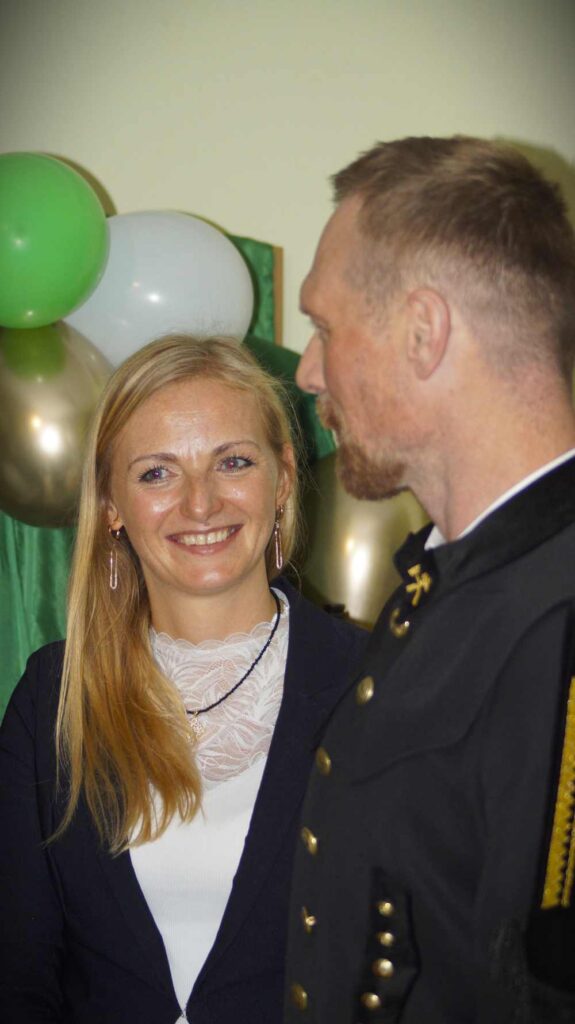 Kobieta z długimi blond włosami uśmiecha się, stojąc obok mężczyzny w ciemnym mundurze na imprezie z zielonymi, białymi i złotymi balonami w tle.