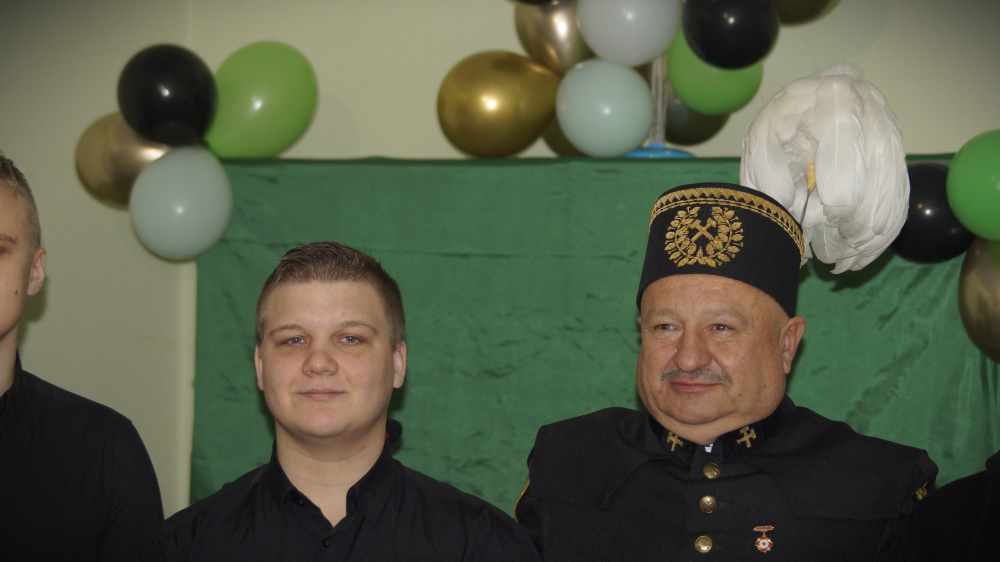 Dwóch mężczyzn stoi przed zielonym tłem z czarnymi, zielonymi, złotymi i białymi balonami. Jeden z nich ma na sobie ciemny mundur z wysokim kapeluszem ozdobionym emblematem i białym pióropuszem; drugi ma na sobie czarną koszulę.