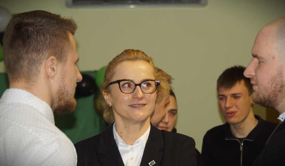 Kobieta w okularach i garniturze stoi wśród czterech mężczyzn zaangażowanych w rozmowę w pomieszczeniu. Wszyscy wydają się rozmawiać lub słuchać, a tło jest delikatnie rozmyte.