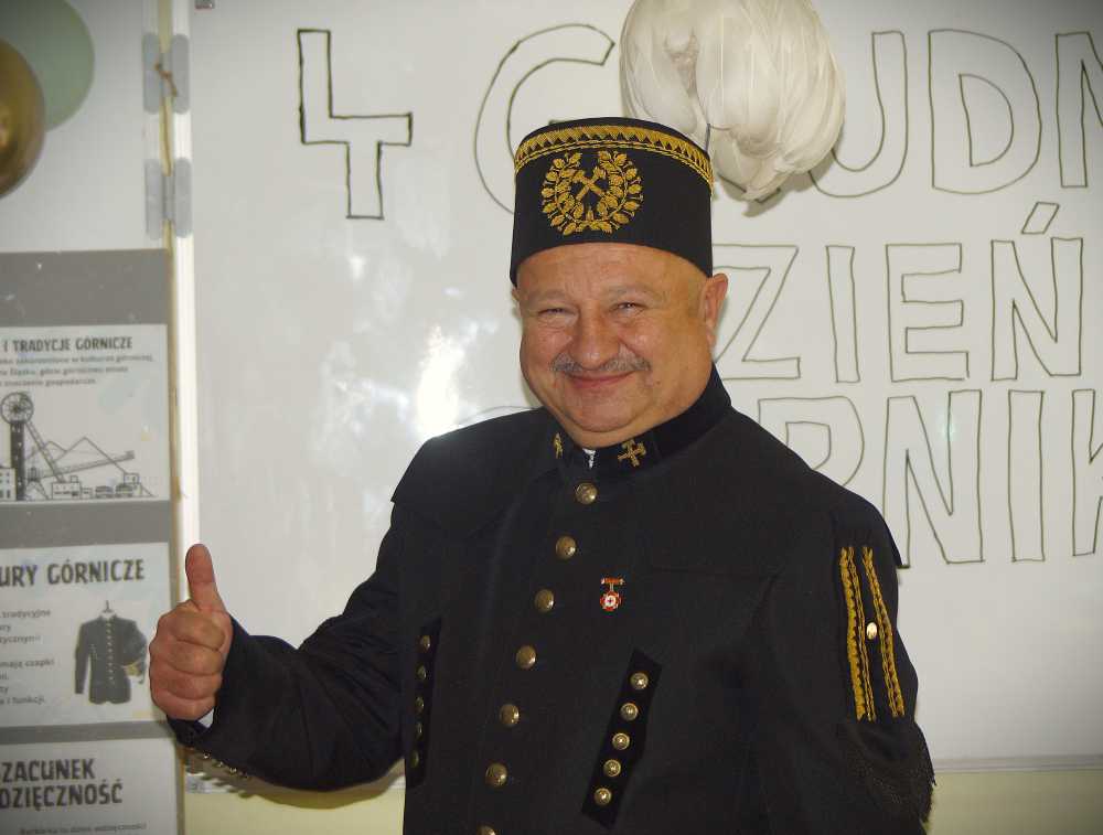 Uśmiechnięty mężczyzna ubrany w tradycyjny czarny mundur górniczy i ozdobną czapkę podnosi kciuk do góry. Za nim na tablicy wyświetlany jest duży, ręcznie rysowany tekst w języku polskim.