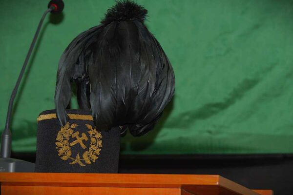 Kapelusz sędziego z czarnym piórem i złotym haftem spoczywa na krześle za drewnianym podium i obok mikrofonu na zielonym tle.