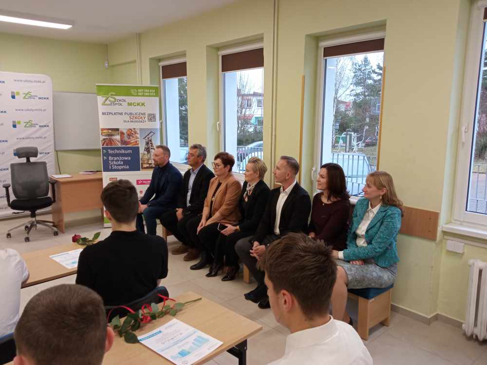 Grupa siedmiu dorosłych w formalnych strojach siedzi w klasie, naprzeciwko kilku uczniów. Na biurkach leżą róże i certyfikaty. Plakaty i okna ustawione są wzdłuż ścian i wygląda na to, że jest to oficjalne wydarzenie lub ceremonia szkolna.