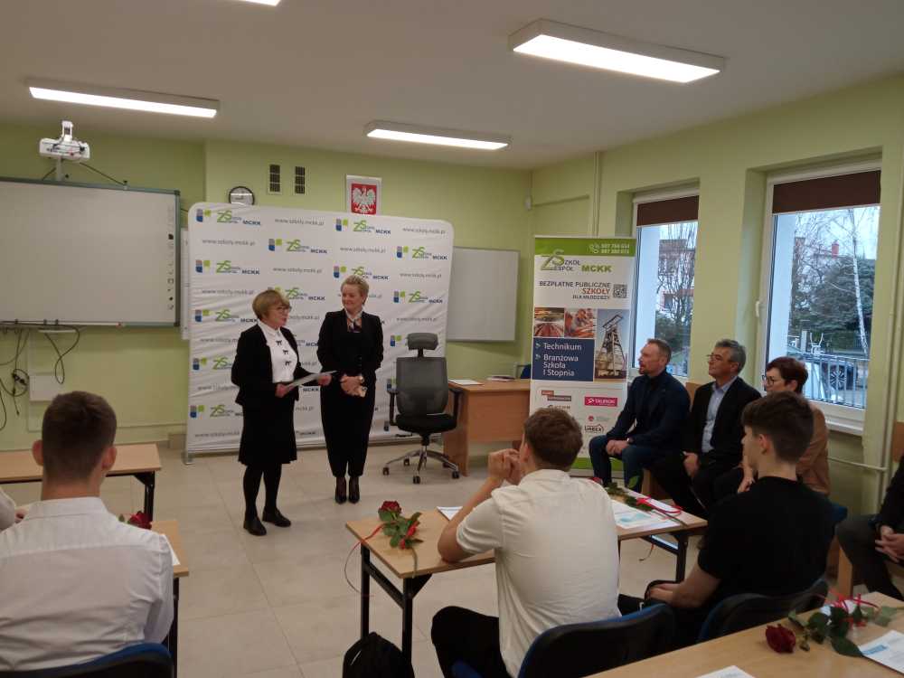 Dwie kobiety stoją z przodu klasy i przemawiają do publiczności. Kilka osób siedzi i słucha, niektórzy trzymają róże. Za mówcami znajdują się banery promocyjne, a okna po prawej stronie pomieszczenia.