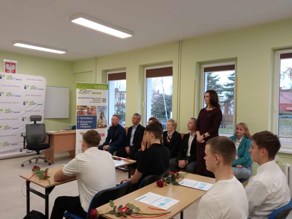 Grupa ludzi, w tym uczniowie siedzący przy biurkach z różami i certyfikatami oraz dorośli siedzący i stojący, zbiera się w klasie z dużymi oknami i banerami promocyjnymi w tle.