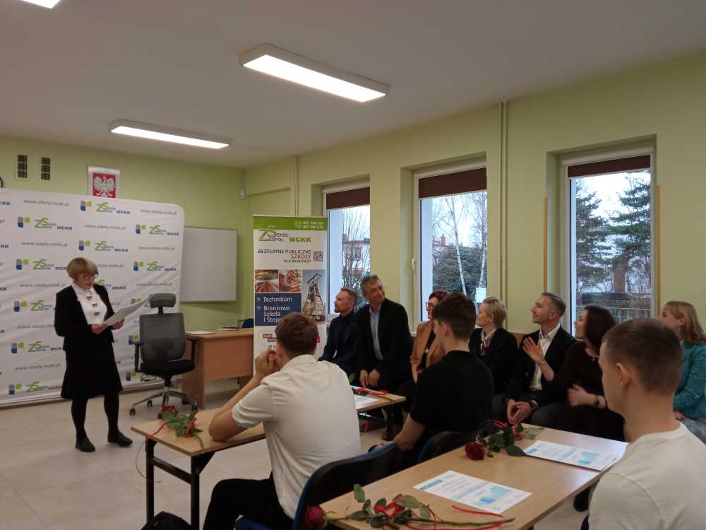 Kobieta przemawia z przodu klasy, podczas gdy kilka osób siedzi i słucha. Niektórzy uczestnicy trzymają certyfikaty i róże. W tle widać banery prezentacyjne i duże okna.