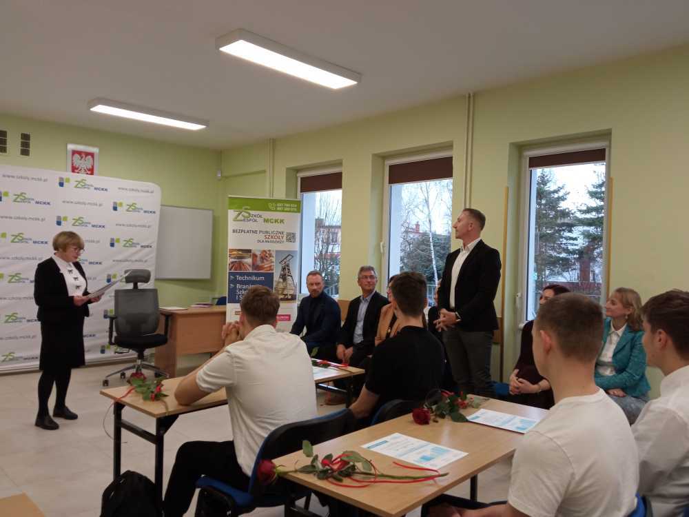 Kobieta stoi z przodu klasy, trzymając dokumenty, zwracając się do siedzącej grupy uczniów i dorosłych. Niektórzy uczniowie mają na biurkach czerwone róże. W tle widoczne są plakaty i okna.