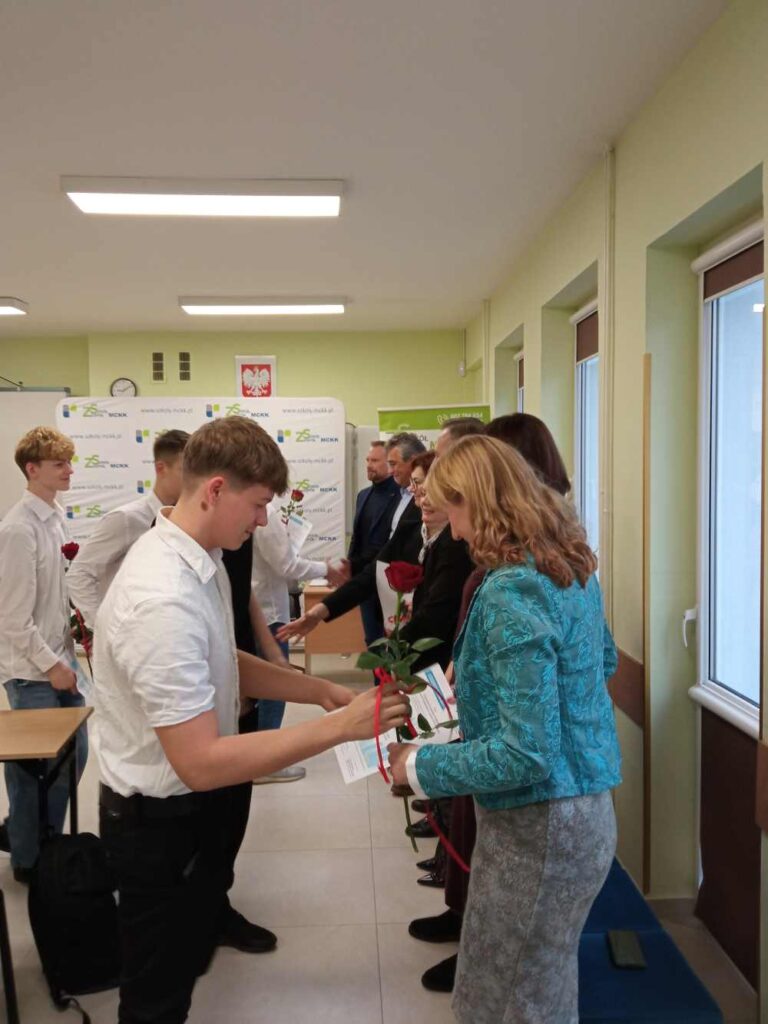 Grupa ludzi stoi w klasie, naprzeciwko siebie w dwóch rzędach. Młodzi mężczyźni wręczają dorosłym róże i papiery, sugerując ceremonię lub rozdanie nagród. Niektórzy dorośli trzymają kwiaty i gazety.