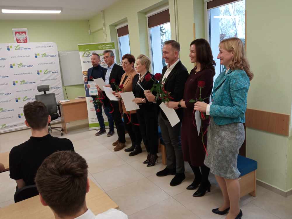 Grupa sześciu dorosłych, z których każdy trzyma pojedynczą czerwoną różę i certyfikat, stoi obok siebie w klasie, podczas gdy siedząca publiczność stoi przed nimi. W tle widoczny jest biały baner i okna.