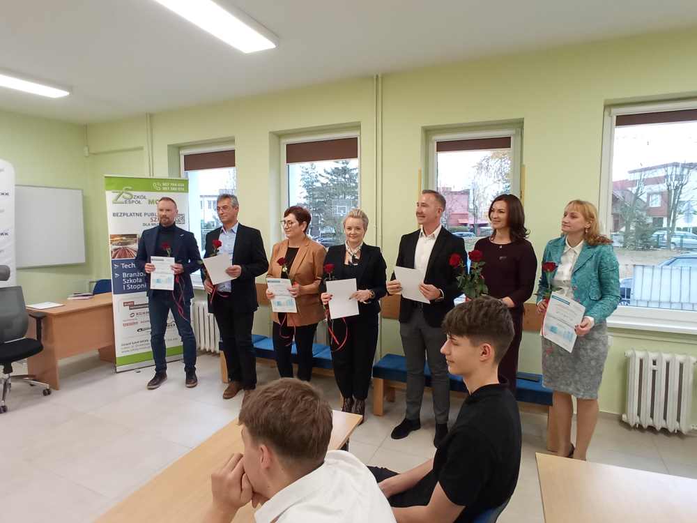 Siedmiu dorosłych stoi w klasie, trzymając certyfikaty i czerwone róże, podczas gdy trzech uczniów siedzi naprzeciwko nich. Grupa stoi w pobliżu okien, a po lewej stronie widoczny jest zwinięty baner.