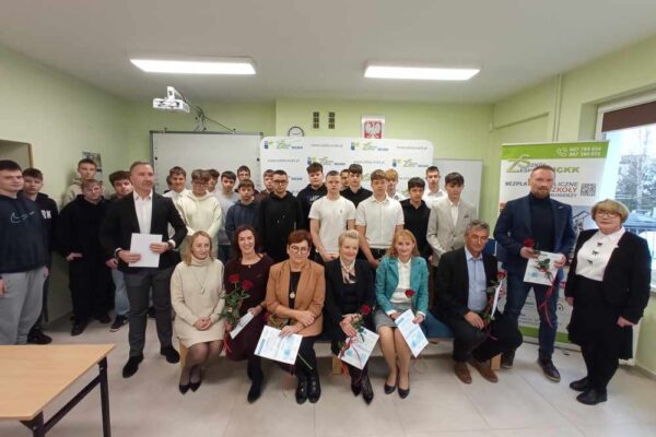 Grupa uczniów i dorosłych pozuje razem w klasie. Dorośli, siedzący z przodu, trzymają certyfikaty i kwiaty, podczas gdy uczniowie stoją za nimi. W tle widać baner i tablice szkolne.