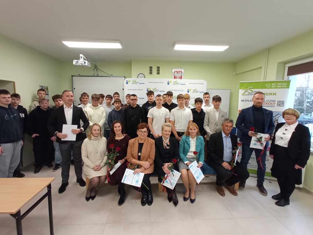 Grupa uczniów i dorosłych pozuje razem w klasie. Dorośli, siedzący z przodu, trzymają certyfikaty i kwiaty, podczas gdy uczniowie stoją za nimi. W tle widać baner i tablice szkolne.