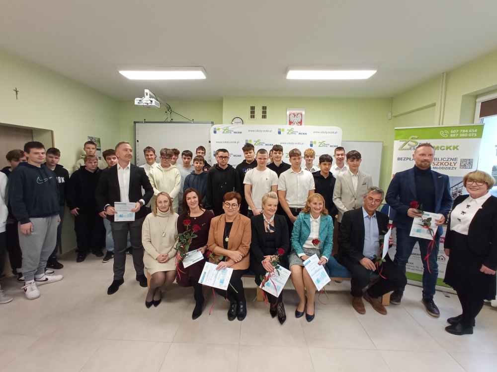 Grupa uczniów i dorosłych pozuje w pomieszczeniu do zdjęcia, niektórzy siedzą trzymając certyfikaty i kwiaty, inni stoją za nimi. W tle widoczny jest baner i ekran projektora.
