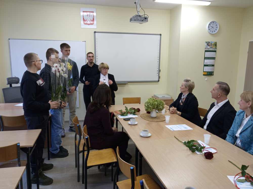 Grupa ludzi siedzi wokół stołu z kwiatami i papierami, podczas gdy cztery stojące osoby, niektóre trzymające kwiaty, stoją przed nimi w klasie z tablicą, zegarem i kalendarzem na ścianie.