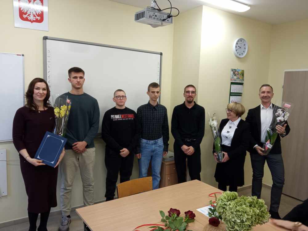 Siedem osób stoi w klasie, niektórzy trzymają kwiaty i prezenty. Na ścianie za nimi znajduje się tablica, projektor i zegar. Na pierwszym planie znajduje się stół z kwiatami i teczką.